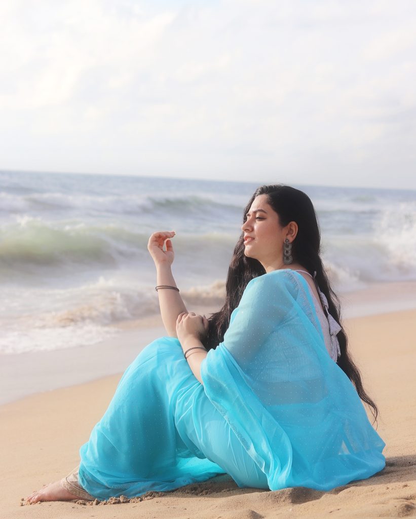 Kanduri Sudha poses gracefully with sandy shoreline and azure blue saree